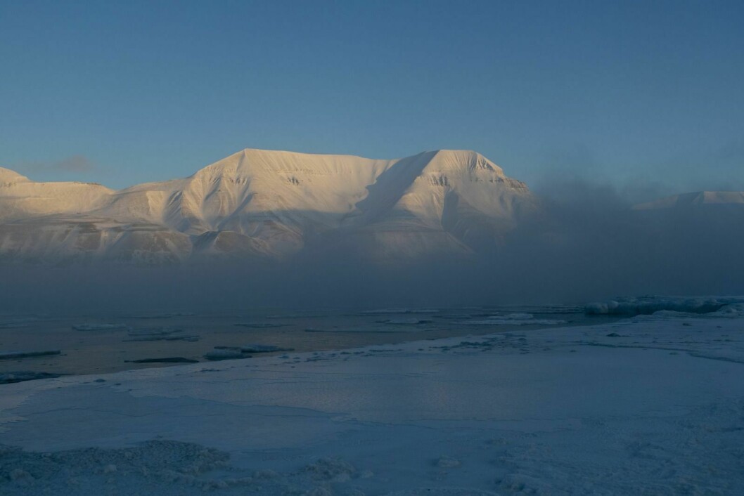 Frostrøyken stiger opp fra Adventfjorden mens de første solstrålene treffer toppen av Hiorthfjellet.